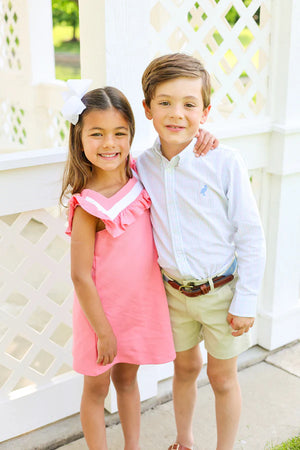Two children stand outdoors, smiling. The girl wears a Girls' Darla Dress by The Beaufort Bonnet Company with a white hair bow; the boy wears a light button-down shirt and khaki shorts.