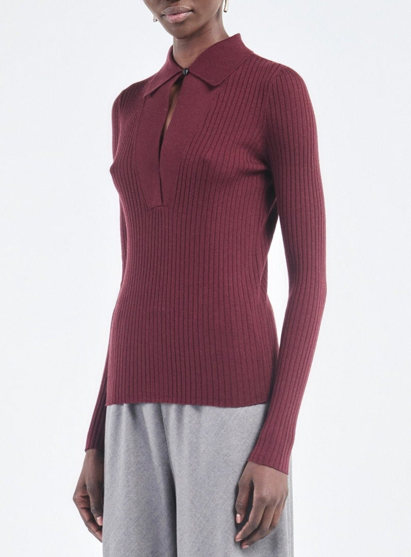 A person wears the Maria McManus Single Button Polo— a slim-fit, ribbed burgundy sweater with a collar and front keyhole—paired with light gray pants.