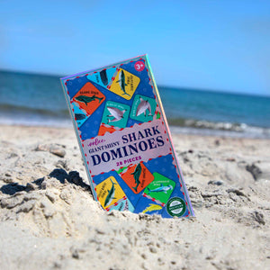 An Eeboo Giant Shiny Shark Dominoes box, a kids’ domino game, stands upright and partly buried in sand on a beach with the sea and blue sky behind it.