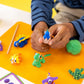A child shapes a blue clay dinosaur from the Floss and Rock Clay & Play - Dinosaur set, surrounded by colorful air dry clay animals and tools on an orange mat, fully immersed in creativity.
