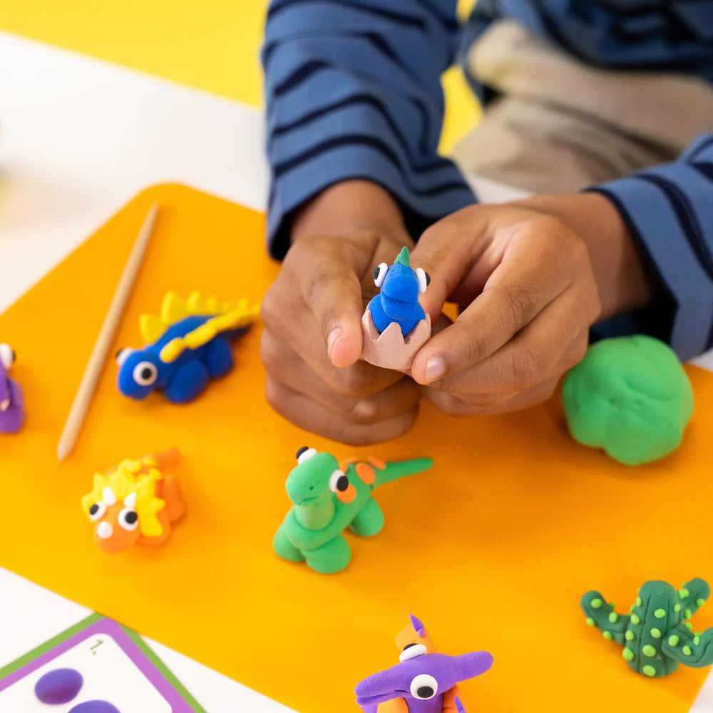 A child shapes a blue clay dinosaur from the Floss and Rock Clay & Play - Dinosaur set, surrounded by colorful air dry clay animals and tools on an orange mat, fully immersed in creativity.
