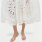 Someone wearing the Simkhai Daila Dress, a white knee-length cotton piece with intricate eyelet embroidery and cut-out patterns, paired with white heeled sandals and standing against a plain background.