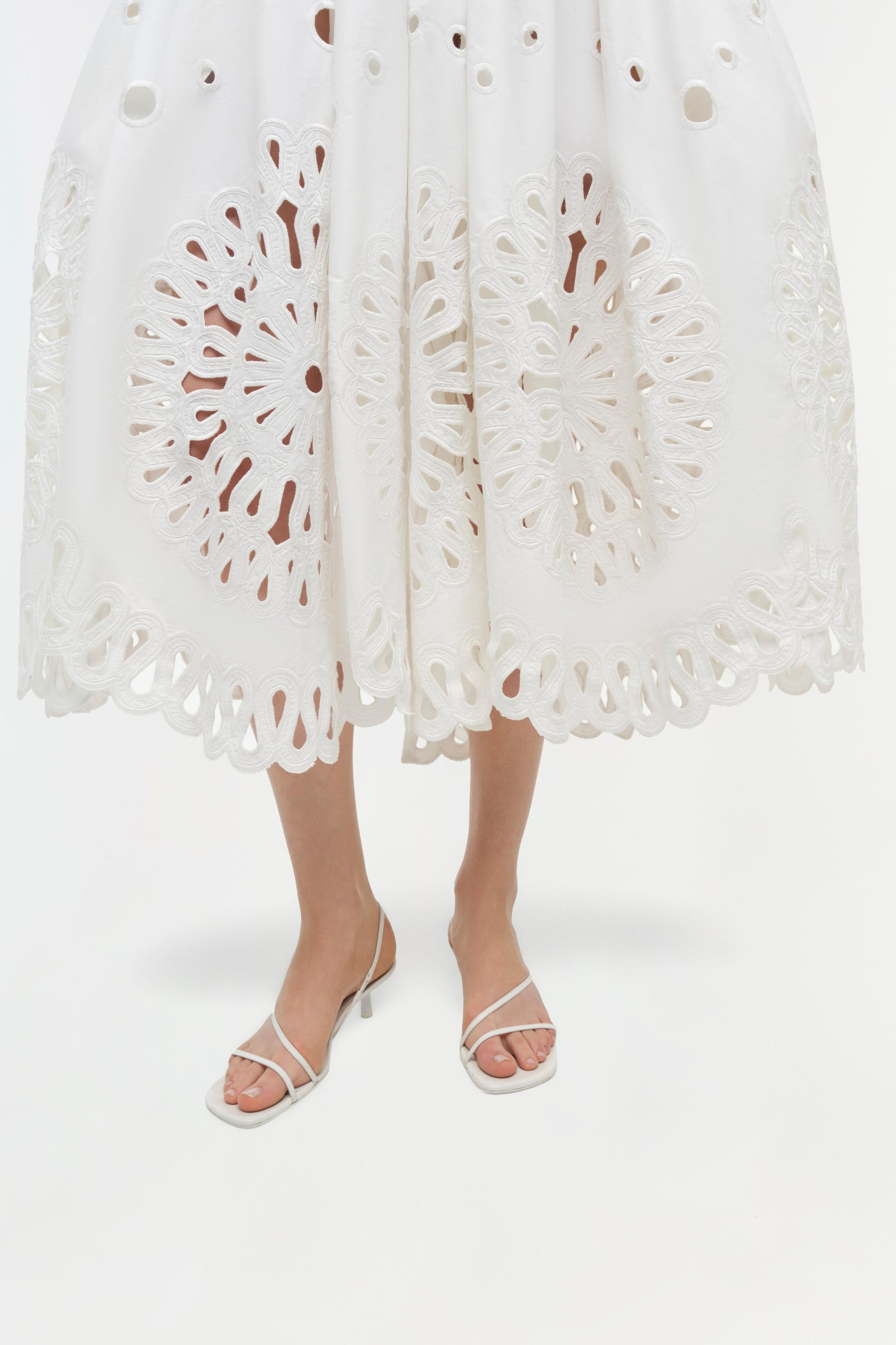 Someone wearing the Simkhai Daila Dress, a white knee-length cotton piece with intricate eyelet embroidery and cut-out patterns, paired with white heeled sandals and standing against a plain background.