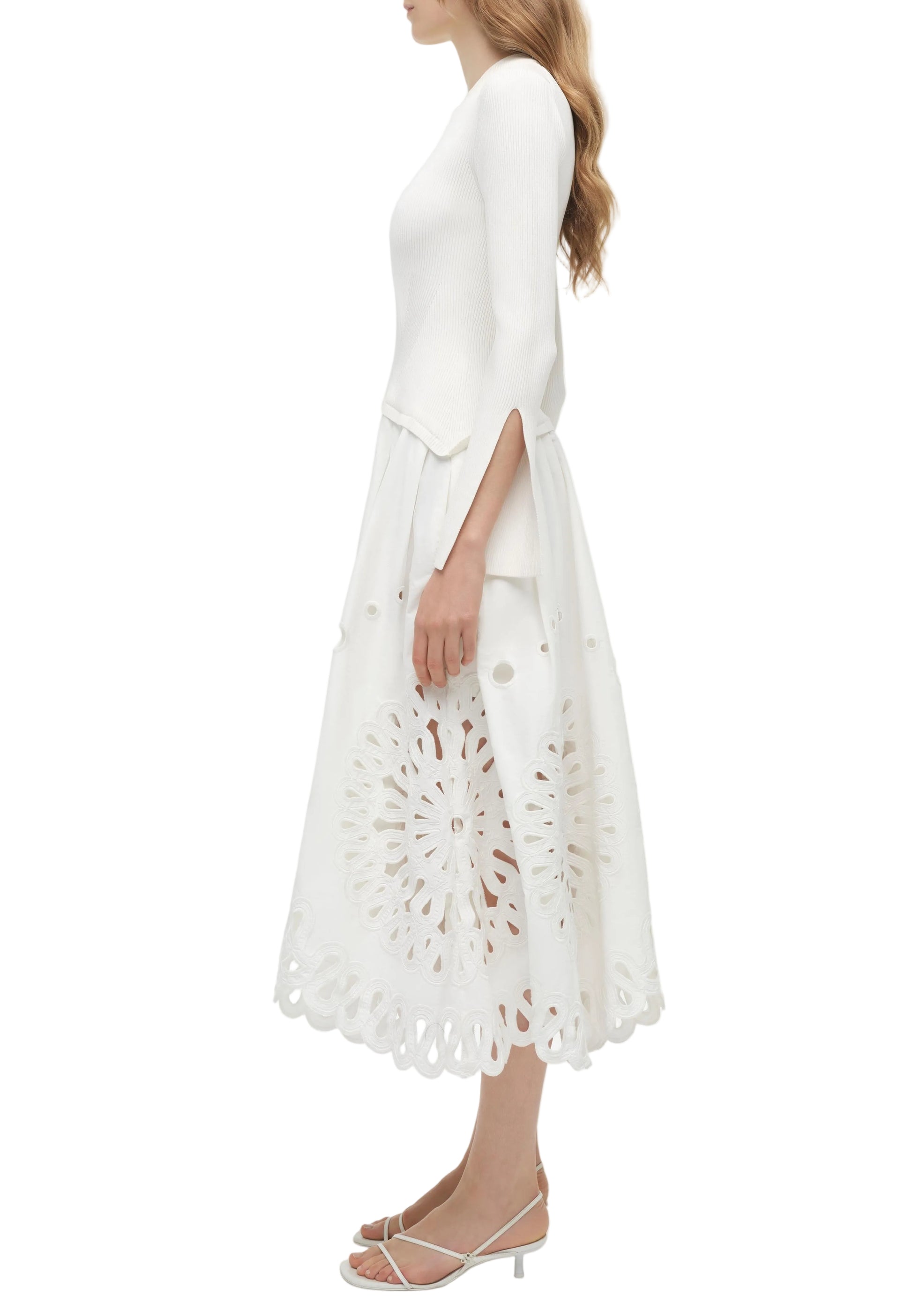 A woman stands in profile wearing the Simkhai Daila Dress—a white long-sleeve piece featuring eyelet embroidery and a scalloped hem—paired with white heeled sandals.