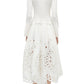 A woman stands facing away, wearing the Simkhai Daila Dress—an eyelet-embroidered cotton piece with long sleeves and a scalloped hem—paired with white sandals.