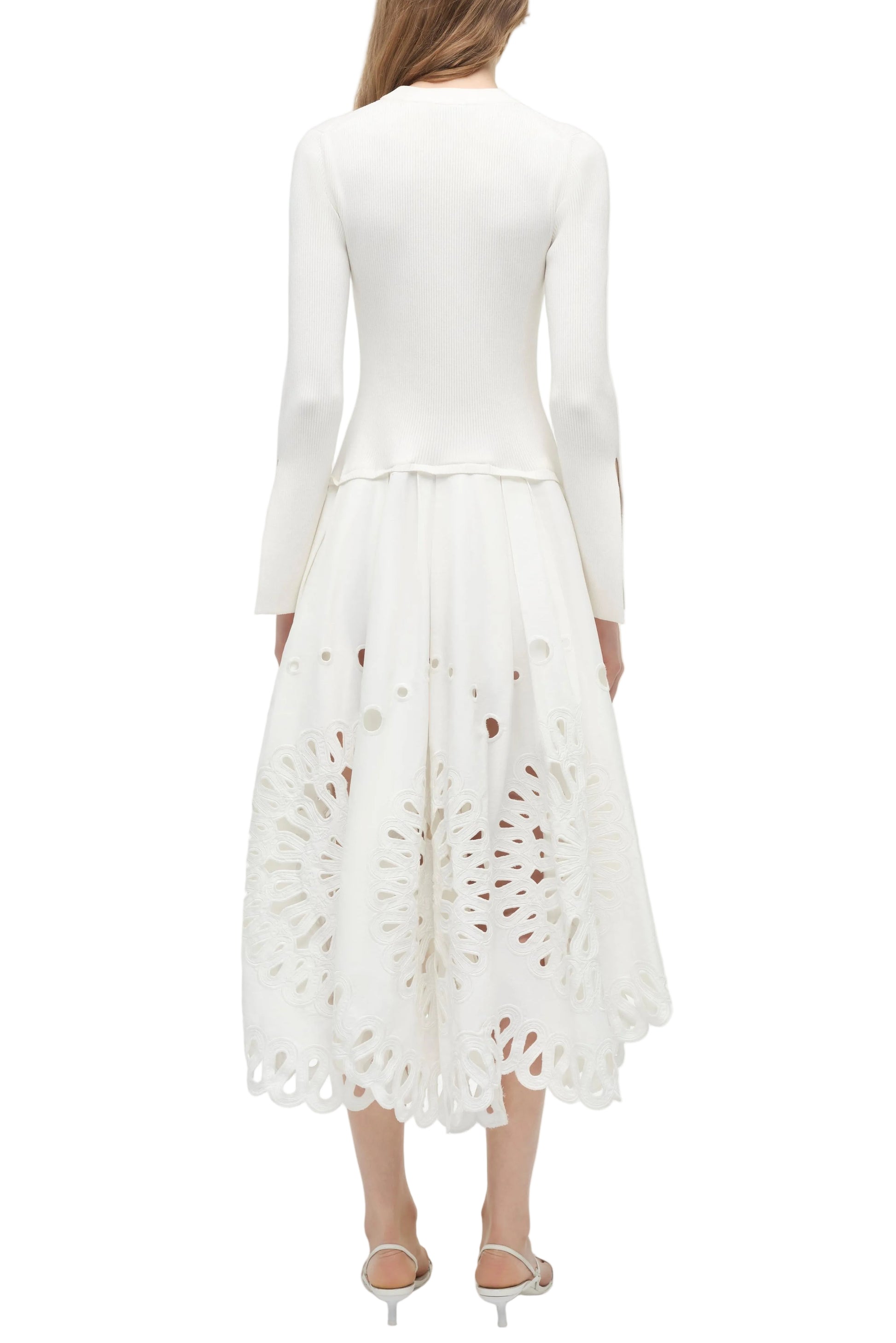 A woman stands facing away, wearing the Simkhai Daila Dress—an eyelet-embroidered cotton piece with long sleeves and a scalloped hem—paired with white sandals.