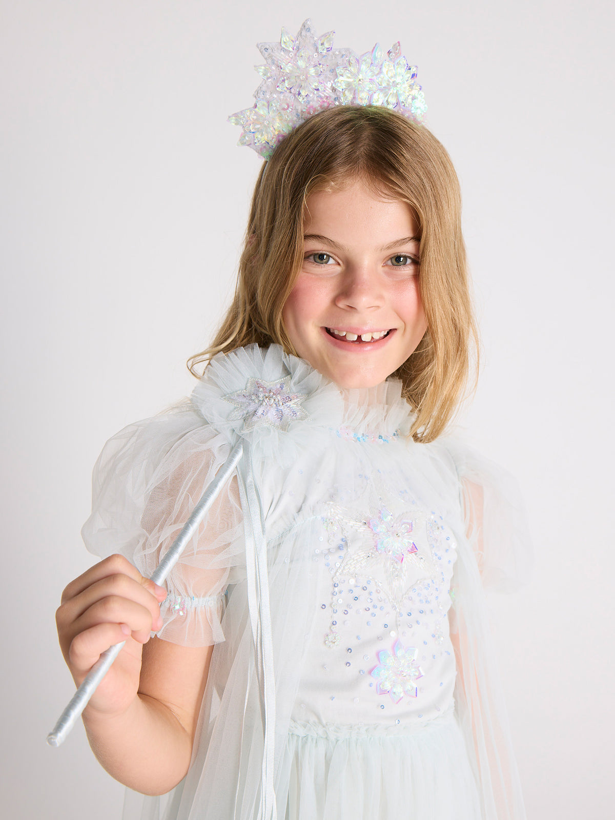 A young girl, dressed in a white, sparkly Snow Queen Tutu Dress from Tutu du Monde, smiles like an icicle princess against a plain light background as she holds the shimmering Snow Queen Wand and wears a matching crown.