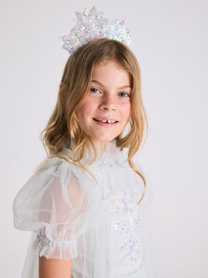 A young girl with long blonde hair is wearing a sparkling Tutu du Monde Snow Queen Tiara and a light blue, glittery princess dress, smiling at the camera against a plain white background.