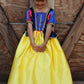 A young girl stands in front of a wooden door, wearing the Great Pretenders Fairytale Princess - Snow White deluxe gown, featuring a blue bodice, yellow skirt, and black shoes crafted with premium quality materials.