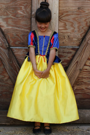 A young girl stands in front of a wooden door, wearing the Great Pretenders Fairytale Princess - Snow White deluxe gown, featuring a blue bodice, yellow skirt, and black shoes crafted with premium quality materials.