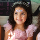 A young girl with face paint and a Great Pretenders Happy Birthday Rhinestone Headband smiles in front of a birthday cake with lit candles.