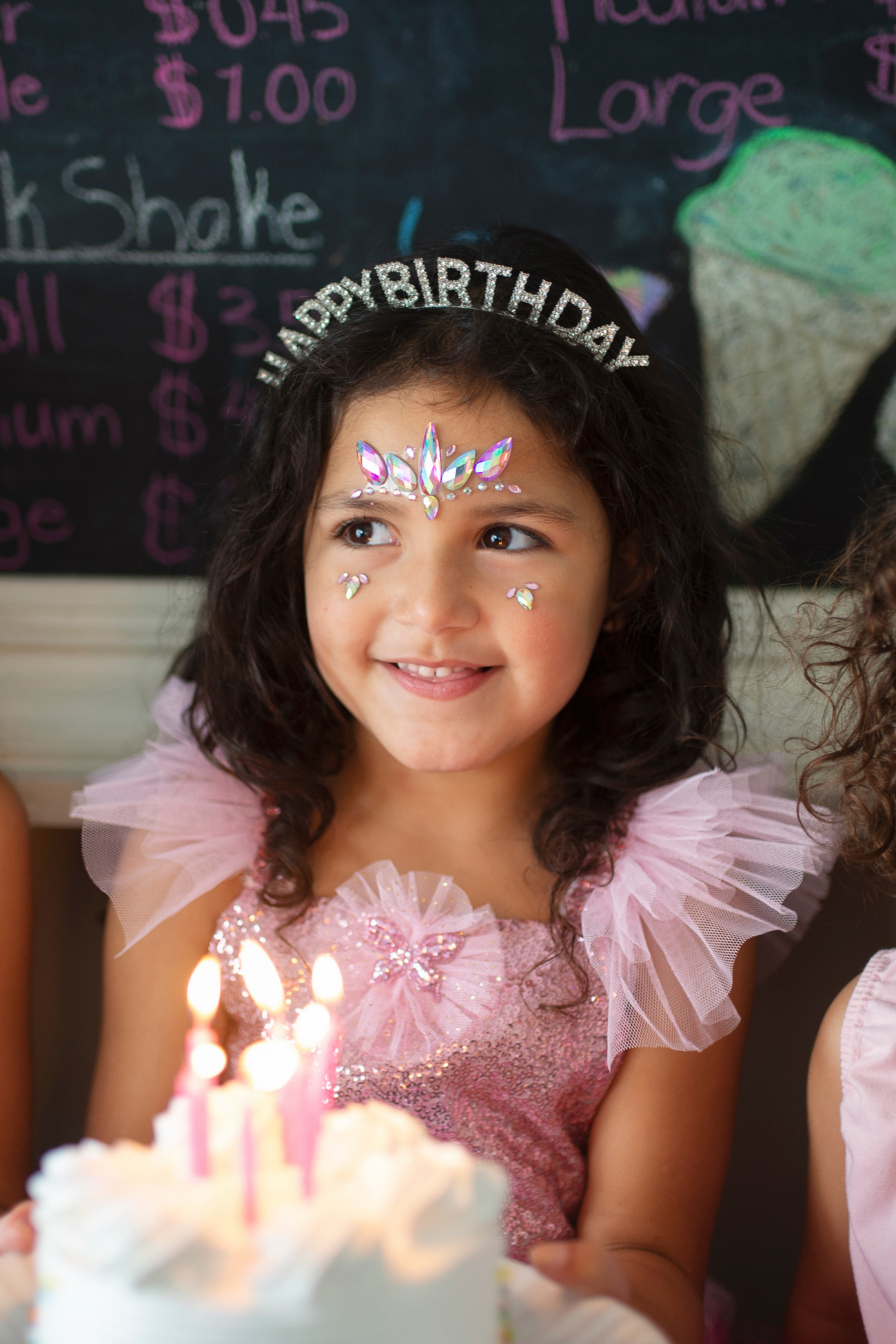 A young girl with face paint and a Great Pretenders Happy Birthday Rhinestone Headband smiles in front of a birthday cake with lit candles.