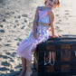 A young girl in the Great Pretenders Bling Bling Mermaid Skirt & Hairclip Set sits on a large wooden chest adorned with beaded necklaces on a sandy beach.
