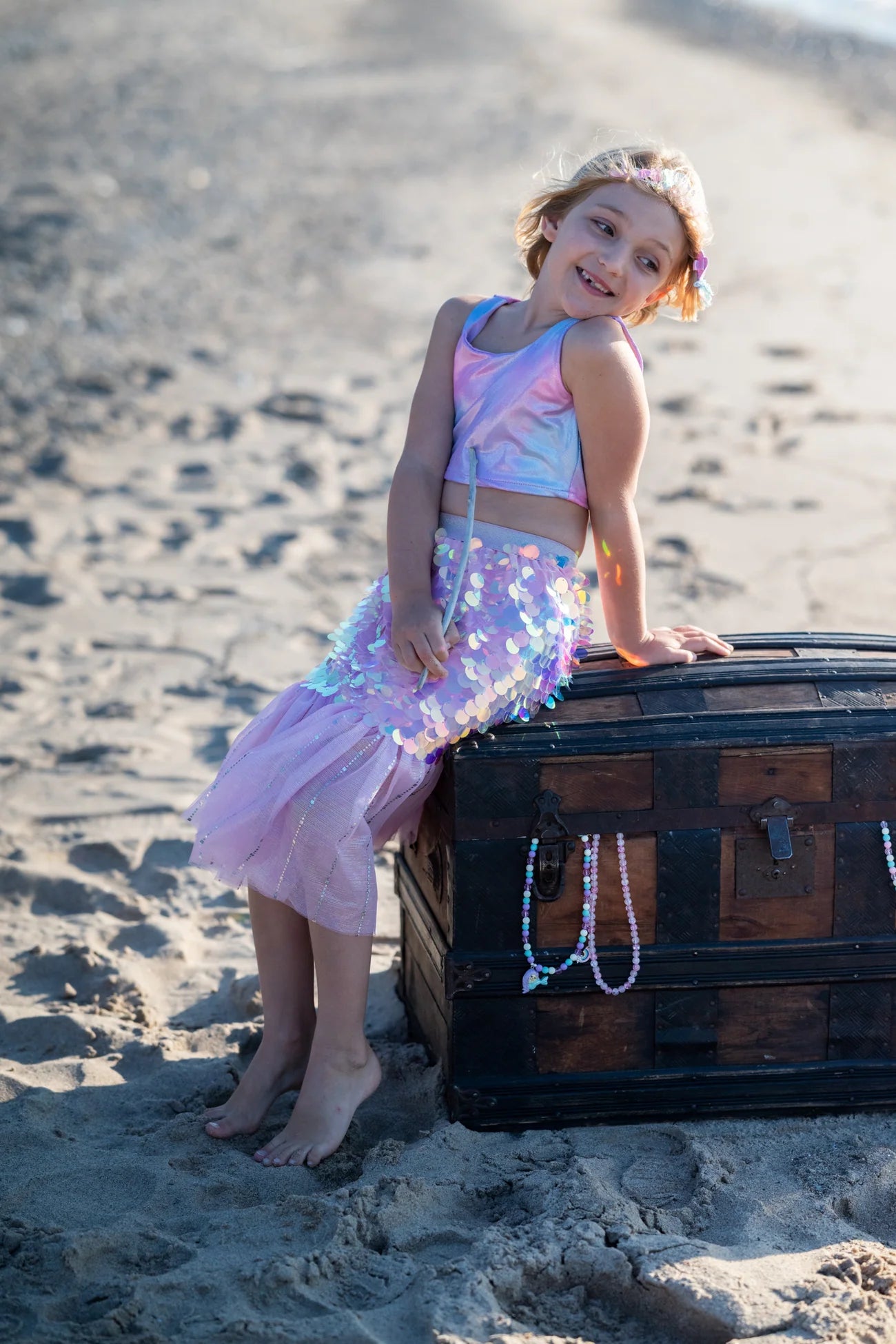 A young girl in the Great Pretenders Bling Bling Mermaid Skirt & Hairclip Set sits on a large wooden chest adorned with beaded necklaces on a sandy beach.
