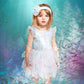 A young girl wearing the Tutu du Monde Baby Girls’ Bebe Mermaid Magic Tutu Dress stands in a dreamy blue and purple fantasy forest illuminated by soft light rays overhead. Delicate starfish motifs shimmer on the skirt, while a dainty headband completes her enchanting look.