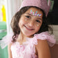 A young girl smiles while wearing a sparkly pink dress, her Great Pretenders Precious Pink Sequins Crown, and face gems, embodying the essence of little princesses and creating lasting childhood memories as she stands indoors.