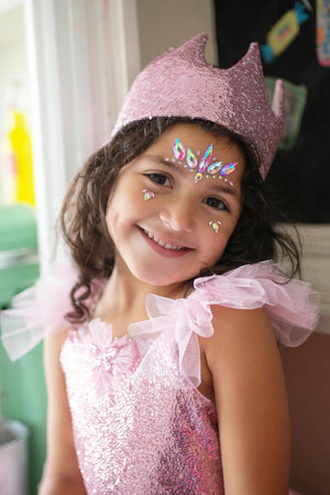 A young girl smiles while wearing a sparkly pink dress, her Great Pretenders Precious Pink Sequins Crown, and face gems, embodying the essence of little princesses and creating lasting childhood memories as she stands indoors.