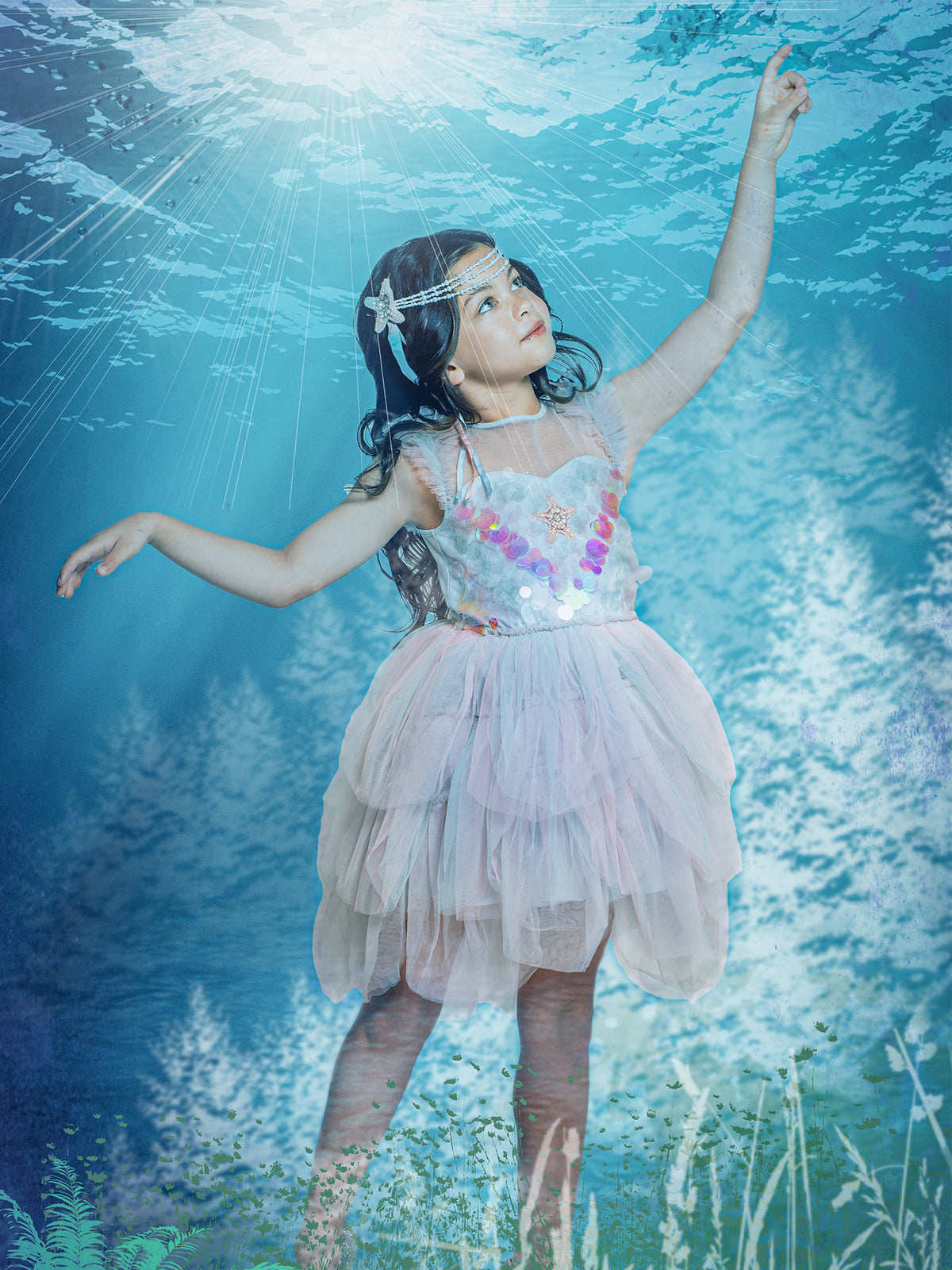 A young girl wearing the Tutu du Monde Girls’ Mermaid Magic Tutu Dress and a matching headband poses underwater, reaching upward toward the light as aquatic plants and sun rays shimmer around her.