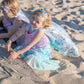 Two young children in vibrant, customizable Mermaid DreamsCape Color-In Capes (ages 4-6) by Great Pretenders play in the sand, one smiling at the camera while the other looks down, partially turned away. Their outfits were colored with washable markers, making each adventure as unique as their imagination.