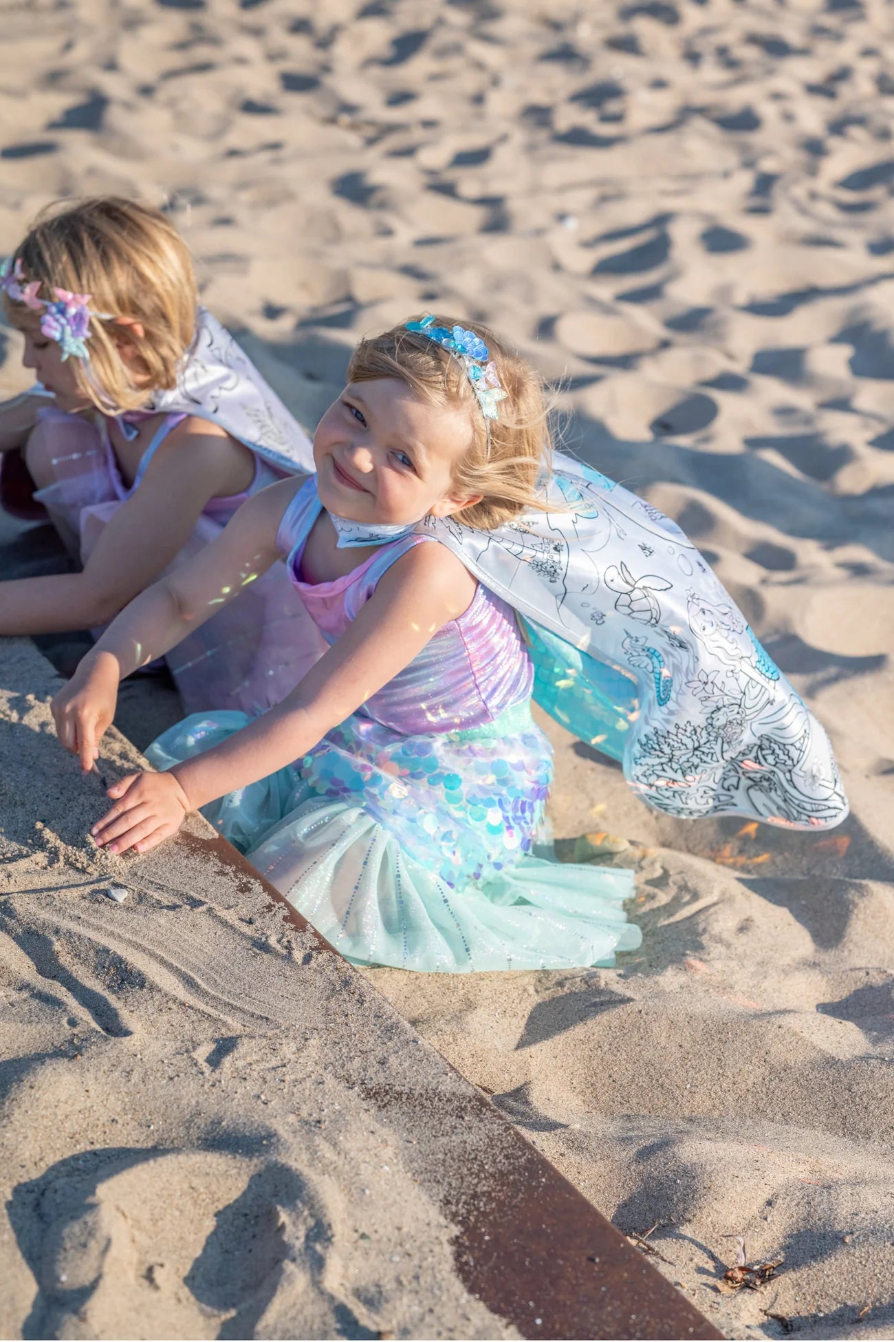 Two young children in vibrant, customizable Mermaid DreamsCape Color-In Capes (ages 4-6) by Great Pretenders play in the sand, one smiling at the camera while the other looks down, partially turned away. Their outfits were colored with washable markers, making each adventure as unique as their imagination.