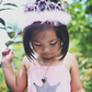 A young child wearing a pink dress with a silver crown design and the Great Pretenders Pink Princess Tiara stands outdoors, looking down.