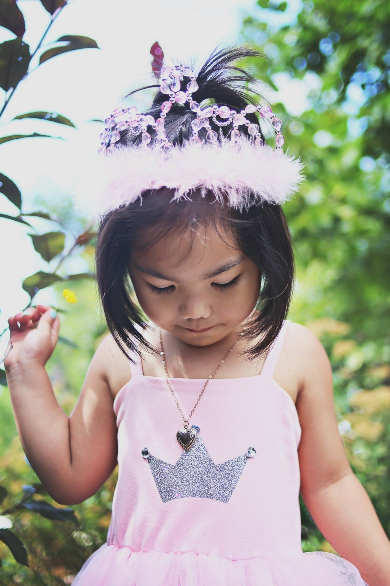 A young child wearing a pink dress with a silver crown design and the Great Pretenders Pink Princess Tiara stands outdoors, looking down.