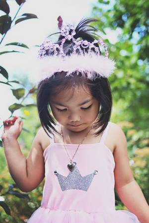 A young child wearing a pink dress with a silver crown design and the Great Pretenders Pink Princess Tiara stands outdoors, looking down.