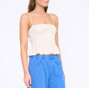 A woman wears the Staud Ayla Linen Tank—a sleeveless, beige cami with a smocked bodice and peplum cropped fit—paired with blue pants against a plain white background.