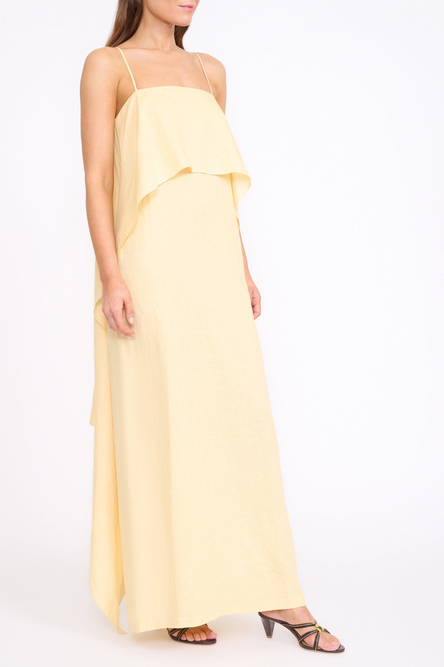 A woman wears the Staud Valencia Linen Dress, a sleeveless light yellow dress with adjustable straps, paired with black strappy sandals, against a plain white background.