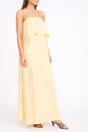 A woman wears the Staud Valencia Linen Dress, a sleeveless light yellow dress with adjustable straps, paired with black strappy sandals, against a plain white background.