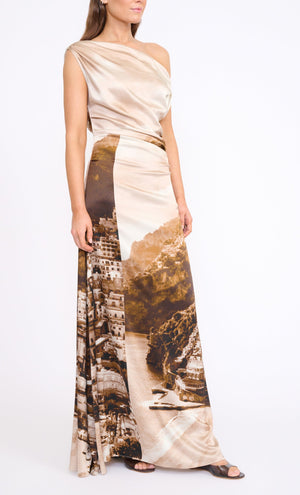 Woman wearing the Staud Phare Silk Maxi Dress, featuring a one-shoulder neckline and a landscape print of buildings and coastline, standing against a plain white background.