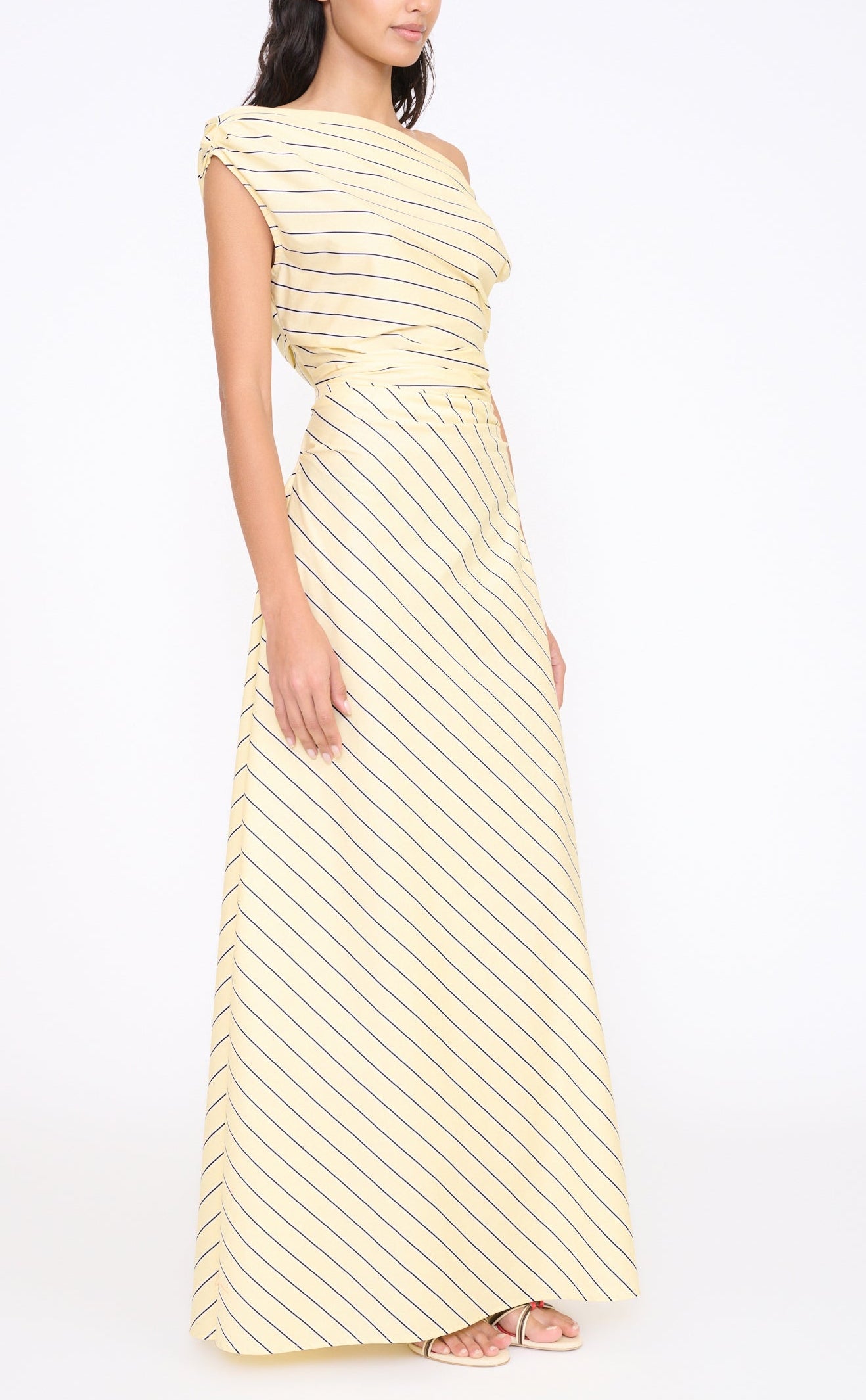 A woman in the Staud Phare Maxi Dress—yellow, off-the-shoulder, with diagonal black stripes and an A-line skirt—paired with open-toe heels, stands against a plain white background.
