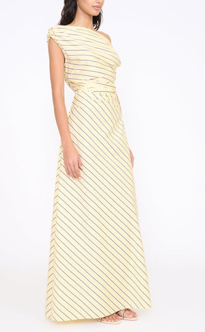 A woman in the Staud Phare Maxi Dress—yellow, off-the-shoulder, with diagonal black stripes and an A-line skirt—paired with open-toe heels, stands against a plain white background.