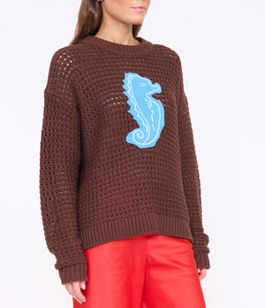 A person wearing the Staud Giada Seahorse Sweater in brown with a blue seahorse design on the front, paired with bright red pants.