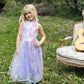 A young girl in the Great Pretenders Ombre Eras Lilac Dress and rain boots stands outdoors on grass beside a floral armchair with a guitar, surrounded by greenery.