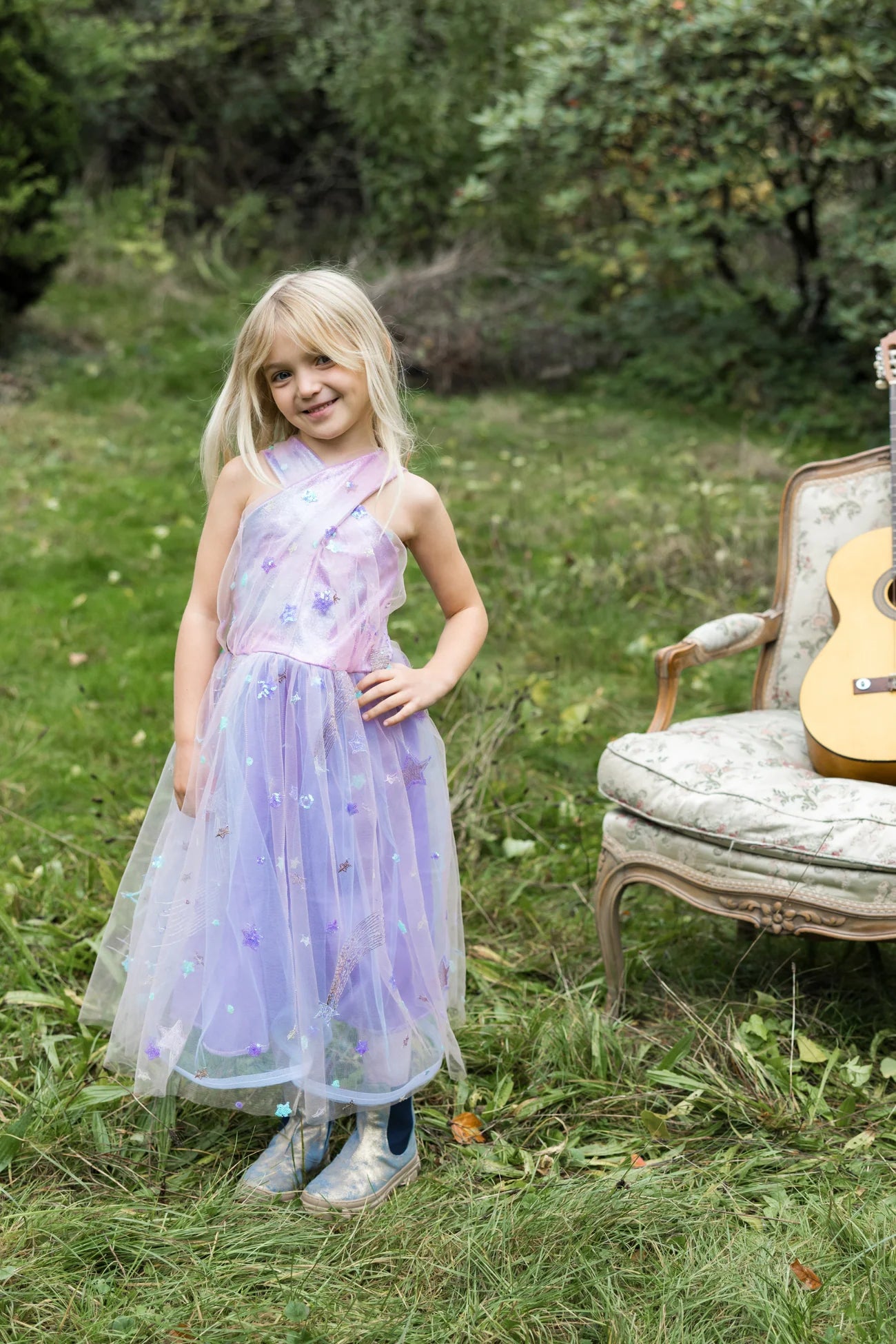 A young girl in the Great Pretenders Ombre Eras Lilac Dress and rain boots stands outdoors on grass beside a floral armchair with a guitar, surrounded by greenery.