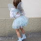 A child dressed in the Great Pretenders Iridescent Celestial Fairy Wings, Tutu, and holding a matching wand (size 4-6Y) stands on a sidewalk, facing away from the camera.