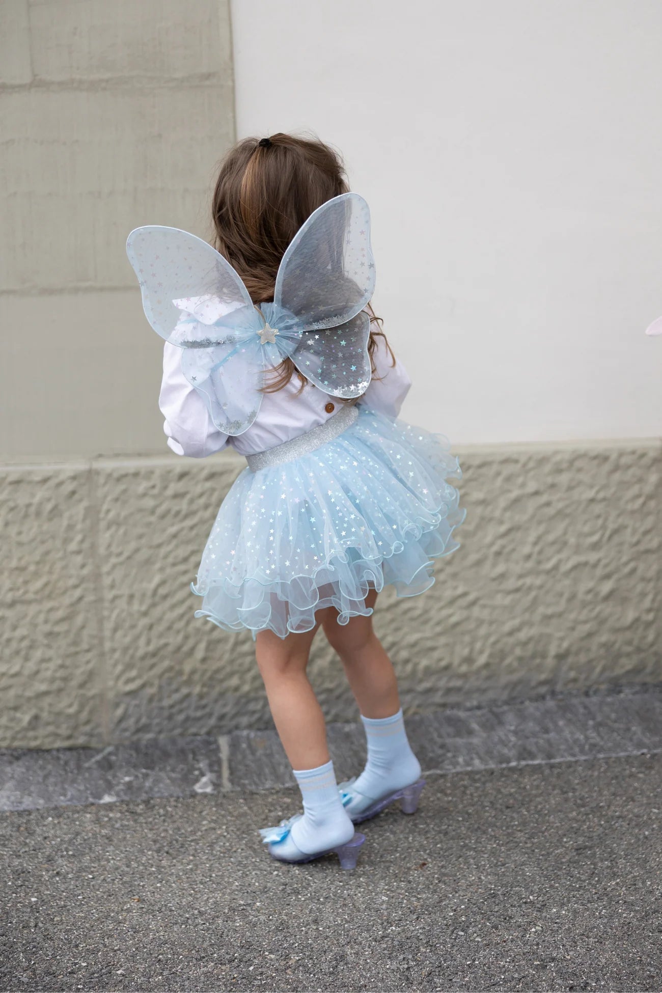A child dressed in the Great Pretenders Iridescent Celestial Fairy Wings, Tutu, and holding a matching wand (size 4-6Y) stands on a sidewalk, facing away from the camera.