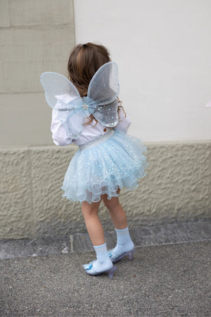A child dressed in the Great Pretenders Iridescent Celestial Fairy Wings, Tutu, and holding a matching wand (size 4-6Y) stands on a sidewalk, facing away from the camera.
