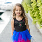 A young girl wearing the Great Pretenders Anna Northern Lights Ballet Tutu Dress smiles at the camera while standing on a walkway near greenery and water.
