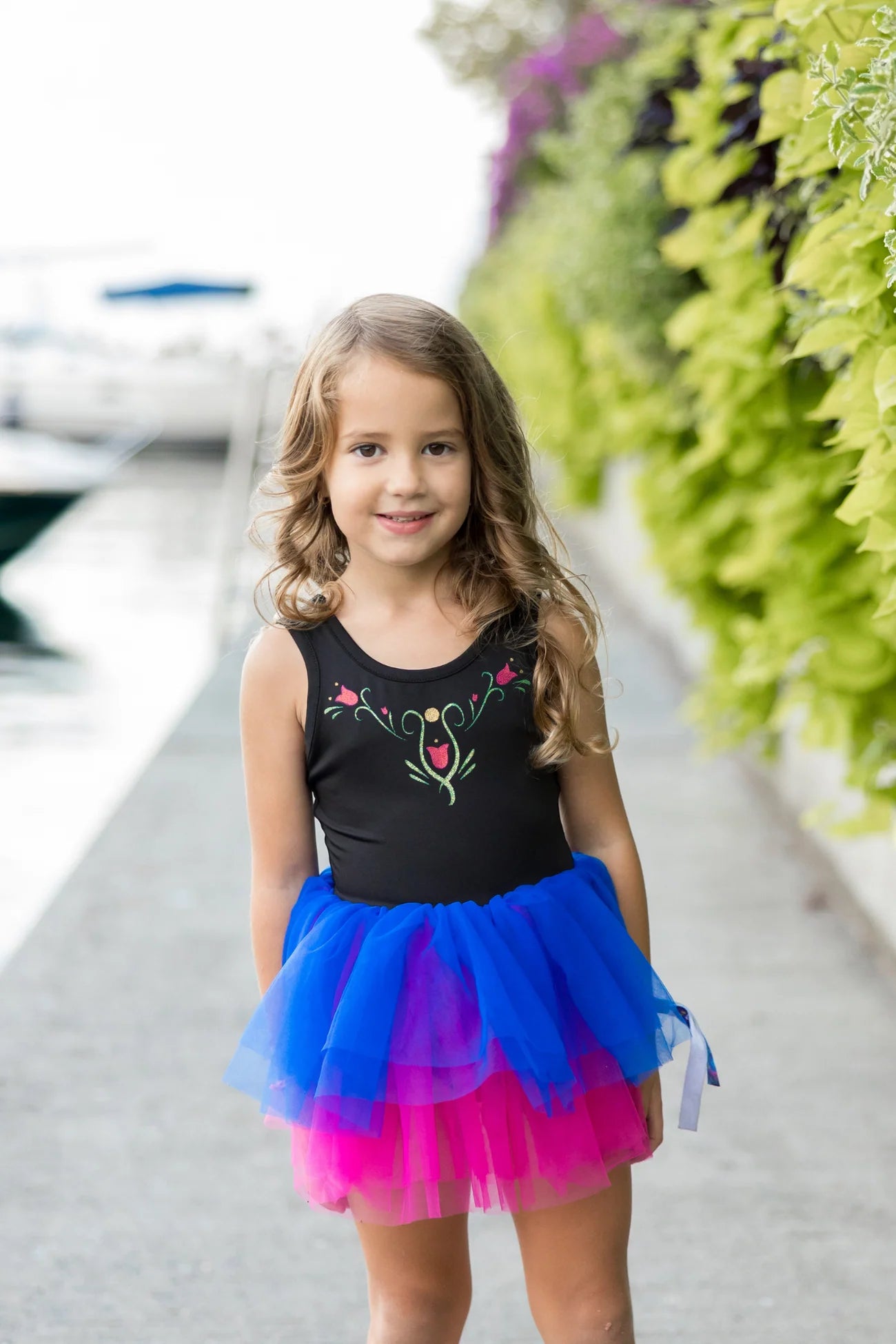 A young girl wearing the Great Pretenders Anna Northern Lights Ballet Tutu Dress smiles at the camera while standing on a walkway near greenery and water.
