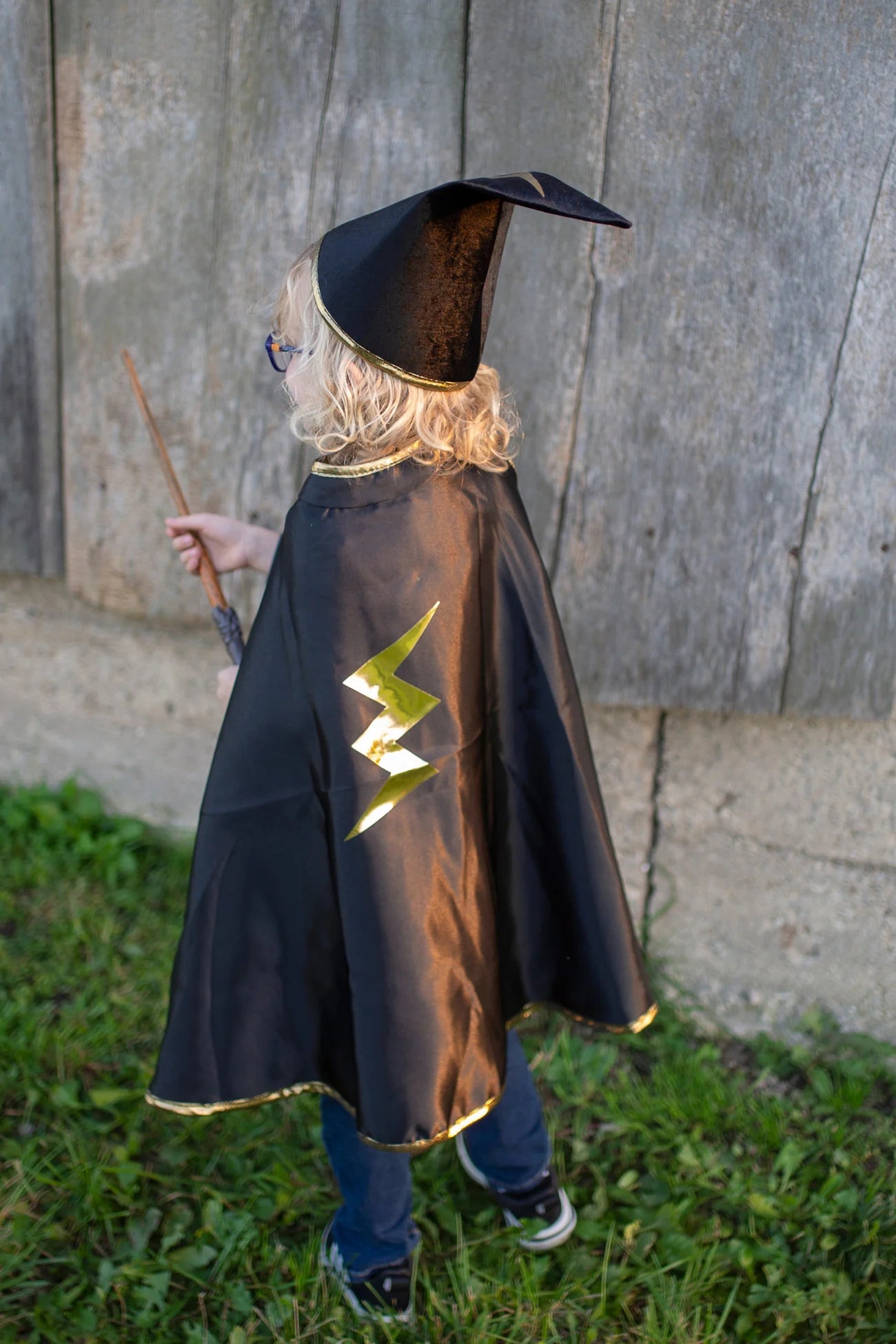 A child dressed in the Great Pretenders Reversible Wizard Cape & Hat stands on grass facing a wooden wall, holding a wand. The outfit features a classic wizard hat and a reversible cape for the perfect spellbinding look.