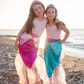 Two shimmering girls in Great Pretenders Mermaid Glimmer Skirt and Tiara on a beach.