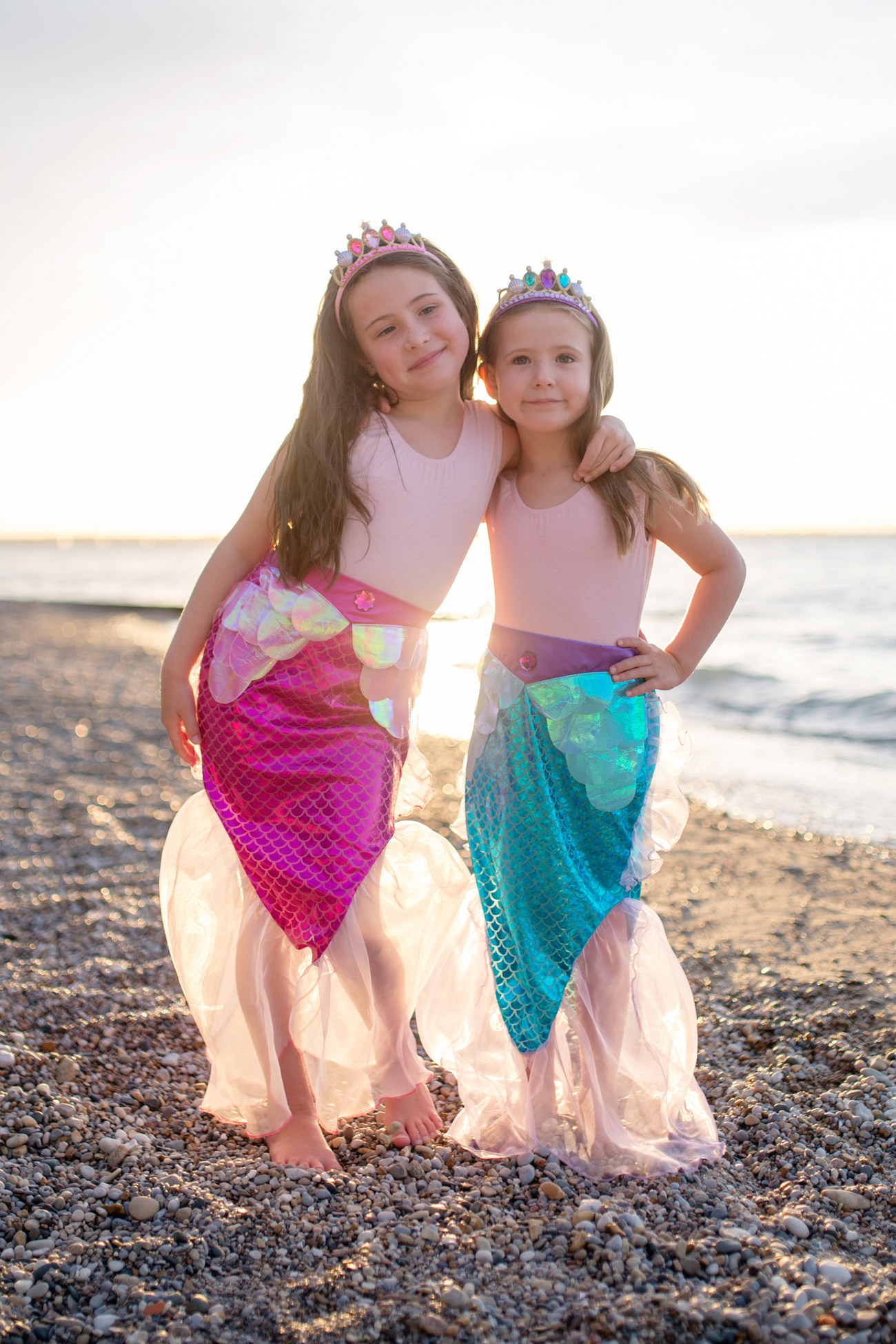 Two shimmering girls in Great Pretenders Mermaid Glimmer Skirt and Tiara on a beach.