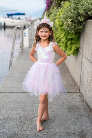 A young girl wears the Great Pretenders Girls' Dreamy Unicorn Dress, barefoot on a waterside walkway with boats and greenery in the background, hands on hips.