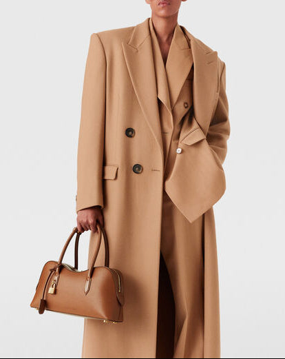 A person in a long camel coat over a matching suit holds the Stella McCartney Ryder Shoulder Bag—a chic, cruelty-free accessory—against a plain background.