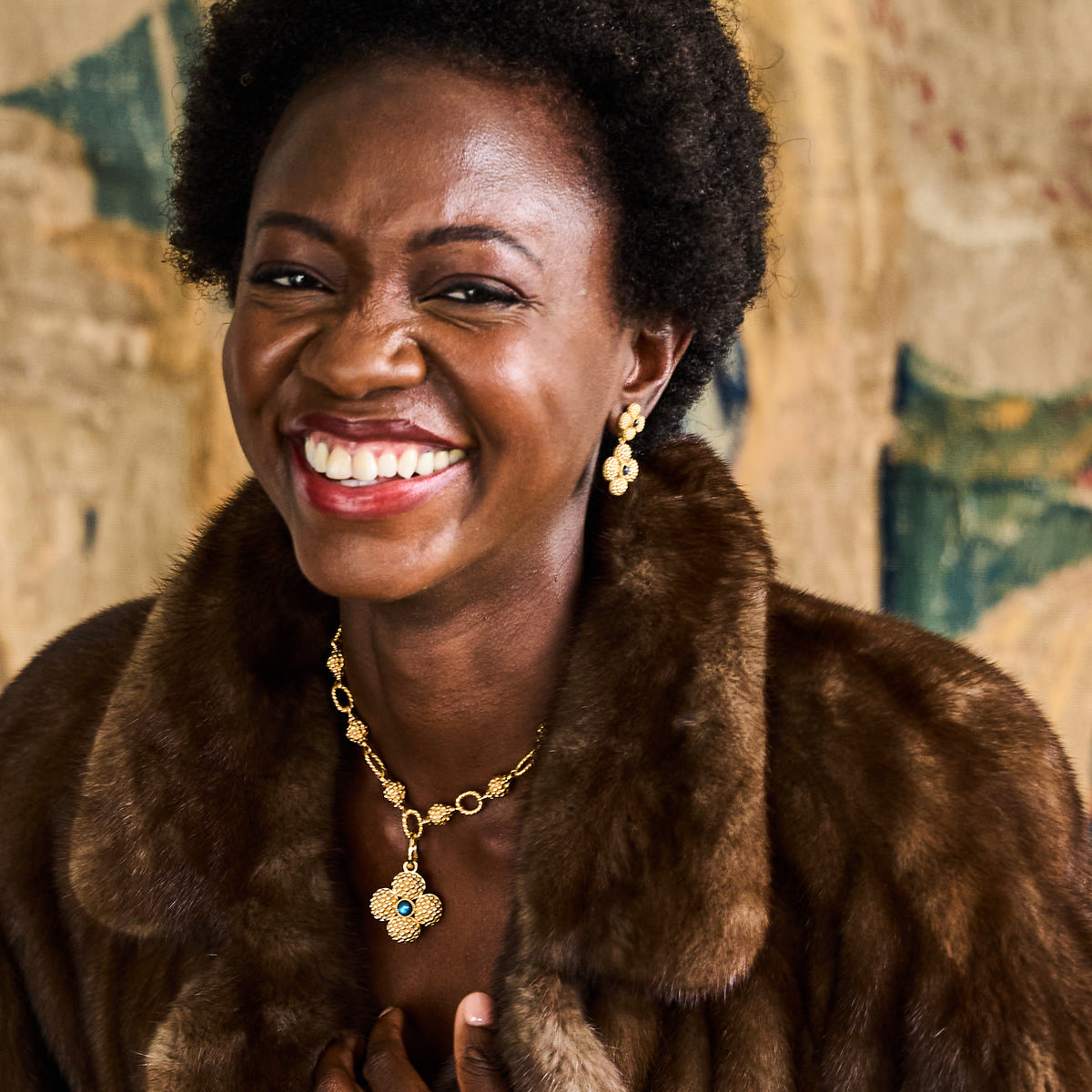 A woman smiles broadly in front of a patterned background, wearing a brown fur coat and Juliska Capucine De Wulf's Berry Clover Double Sided Pendant with matching earrings.
