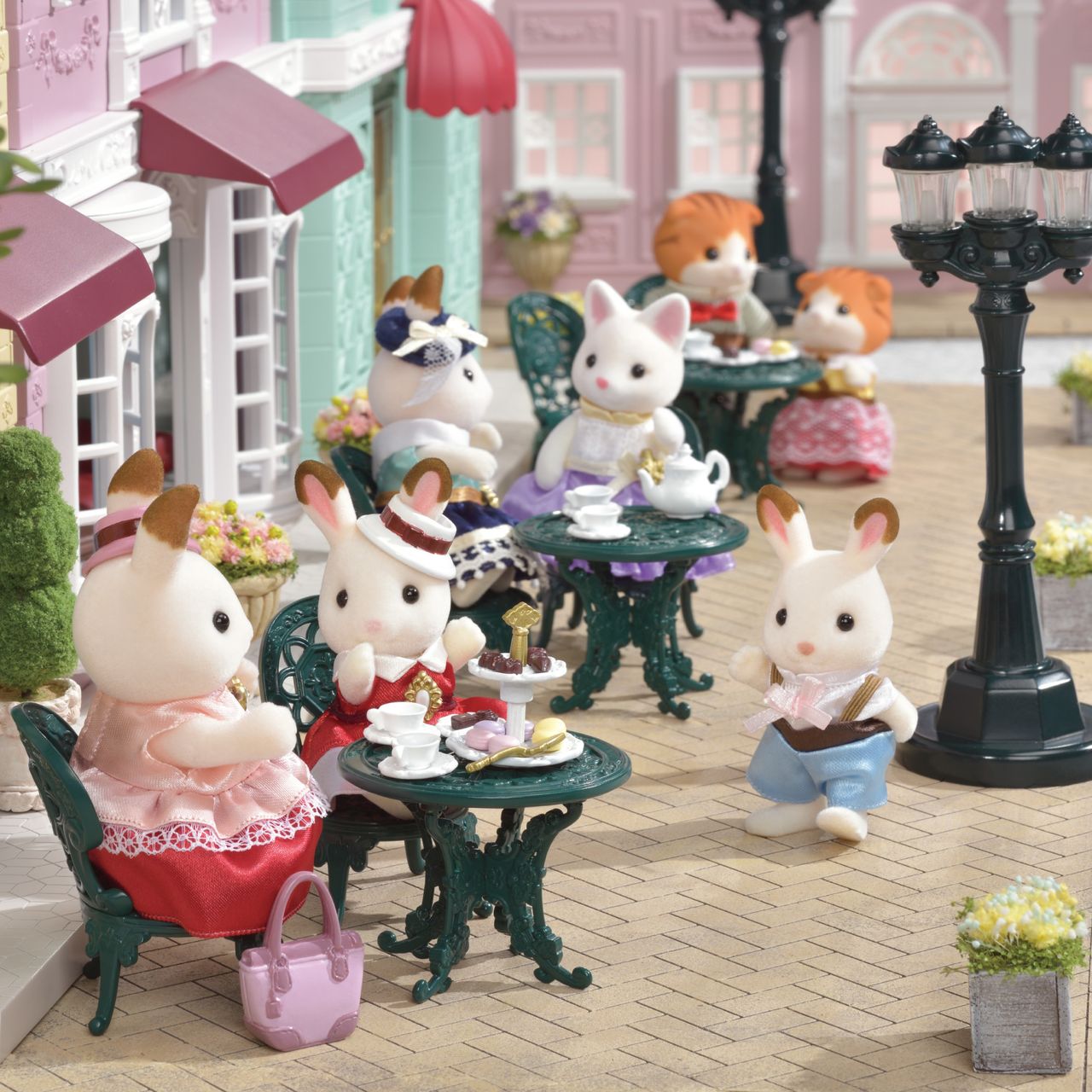 Calico Critters from the Tea and Treats Set gather at outdoor café tables furnished with delightful miniature furniture, sipping tea and enjoying pastries surrounded by charming tea party accessories, while other figures stroll or stand nearby on the cobblestone street.