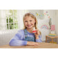 A young girl in a lavender sweater and denim overalls smiles and holds a small Calico Critters Chocolate Rabbit Family figurine while sitting at a wooden table in a bright, decorated bedroom.