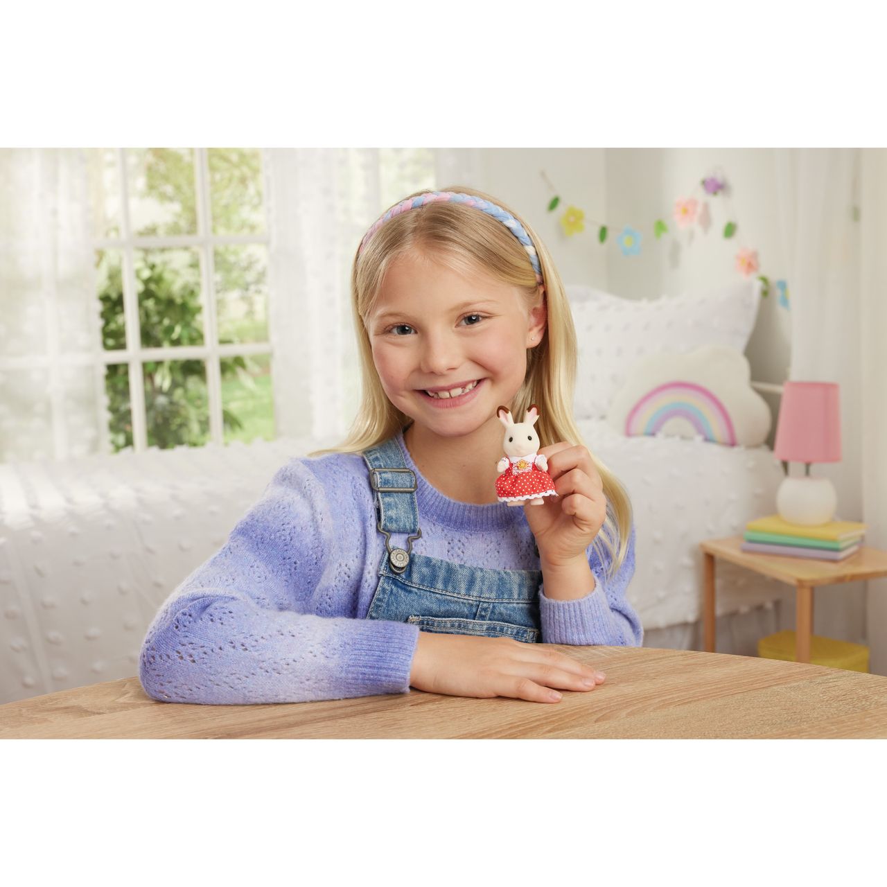 A young girl in a lavender sweater and denim overalls smiles and holds a small Calico Critters Chocolate Rabbit Family figurine while sitting at a wooden table in a bright, decorated bedroom.